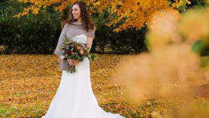 A bride stand outside with ehr flowers infront of autumnal trees