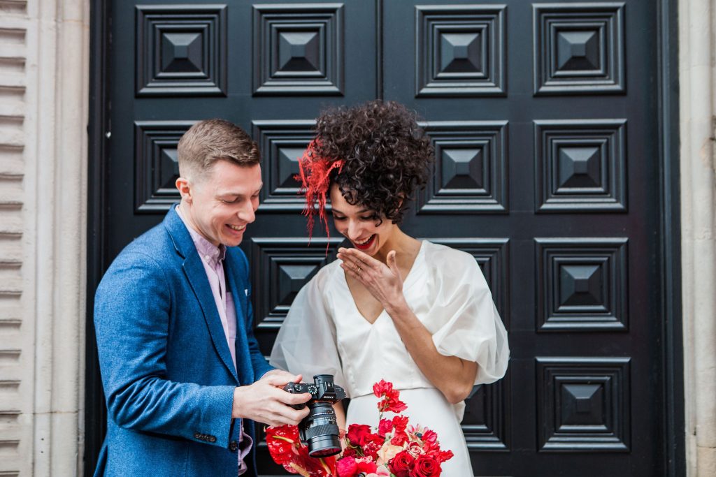 A bride checks out her wedding video shot on the back of a camera the videographer holds for her to see, she is wearing a white dress with her hair up and a red flower in it, the videographer has a smart blue blazer on and they are standing in a doorway
