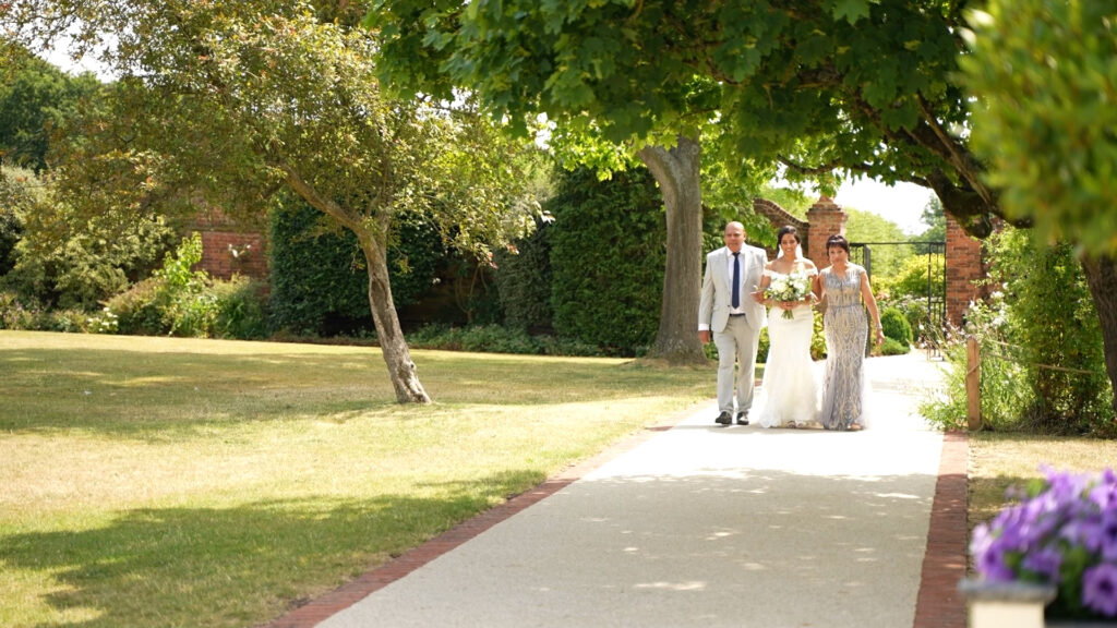 A bride and her parents walk down an long outdoor path through the grounds of a large garden as they prepare to enter her wedding ceremony