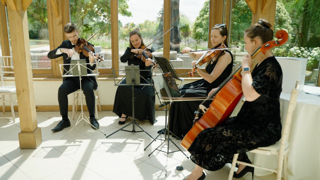 A string quartet sit and play together in an orangery room for the entrance of the bride