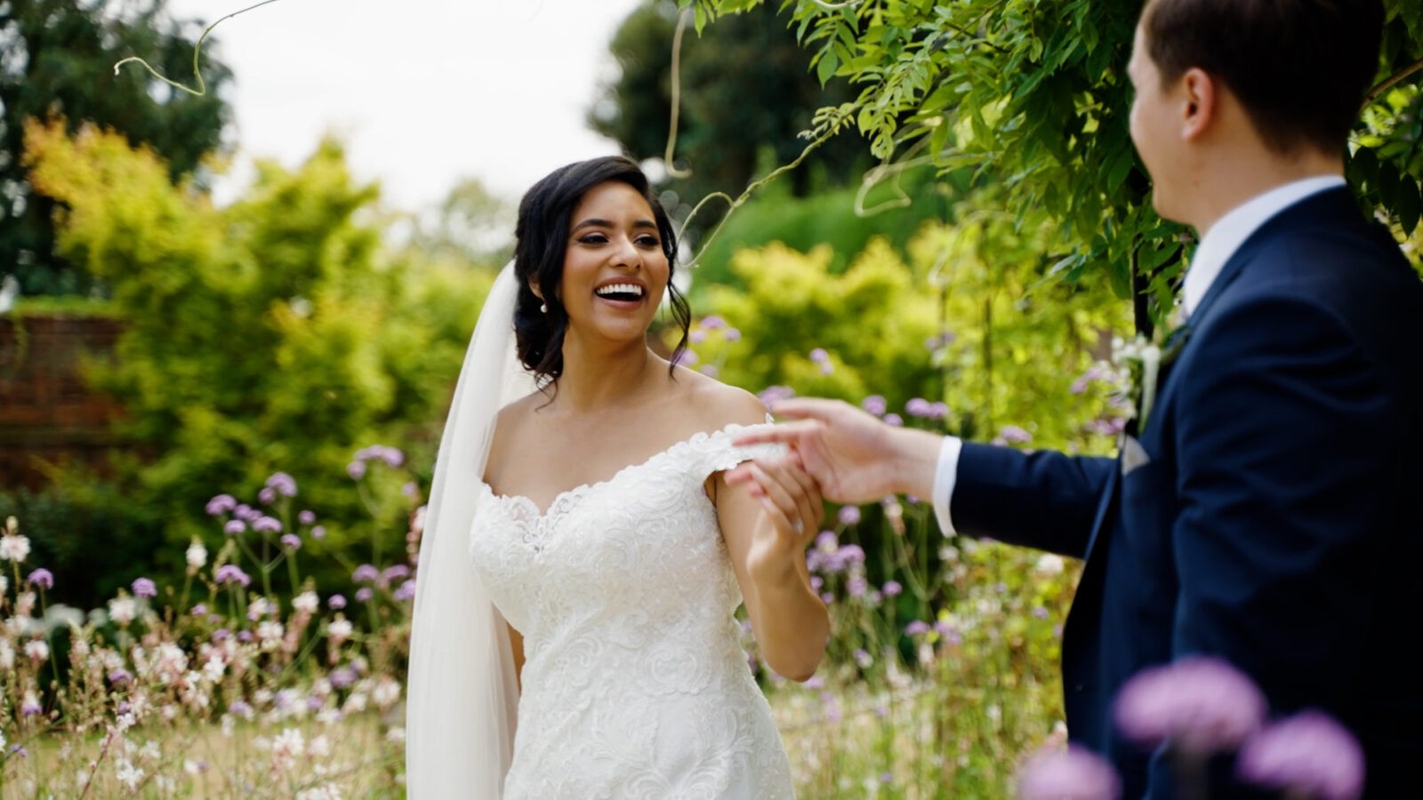 A South African bride stands holding her husbands hand laughing in a flower garden while wearing her white wedding dress and veil, she looks very happy
