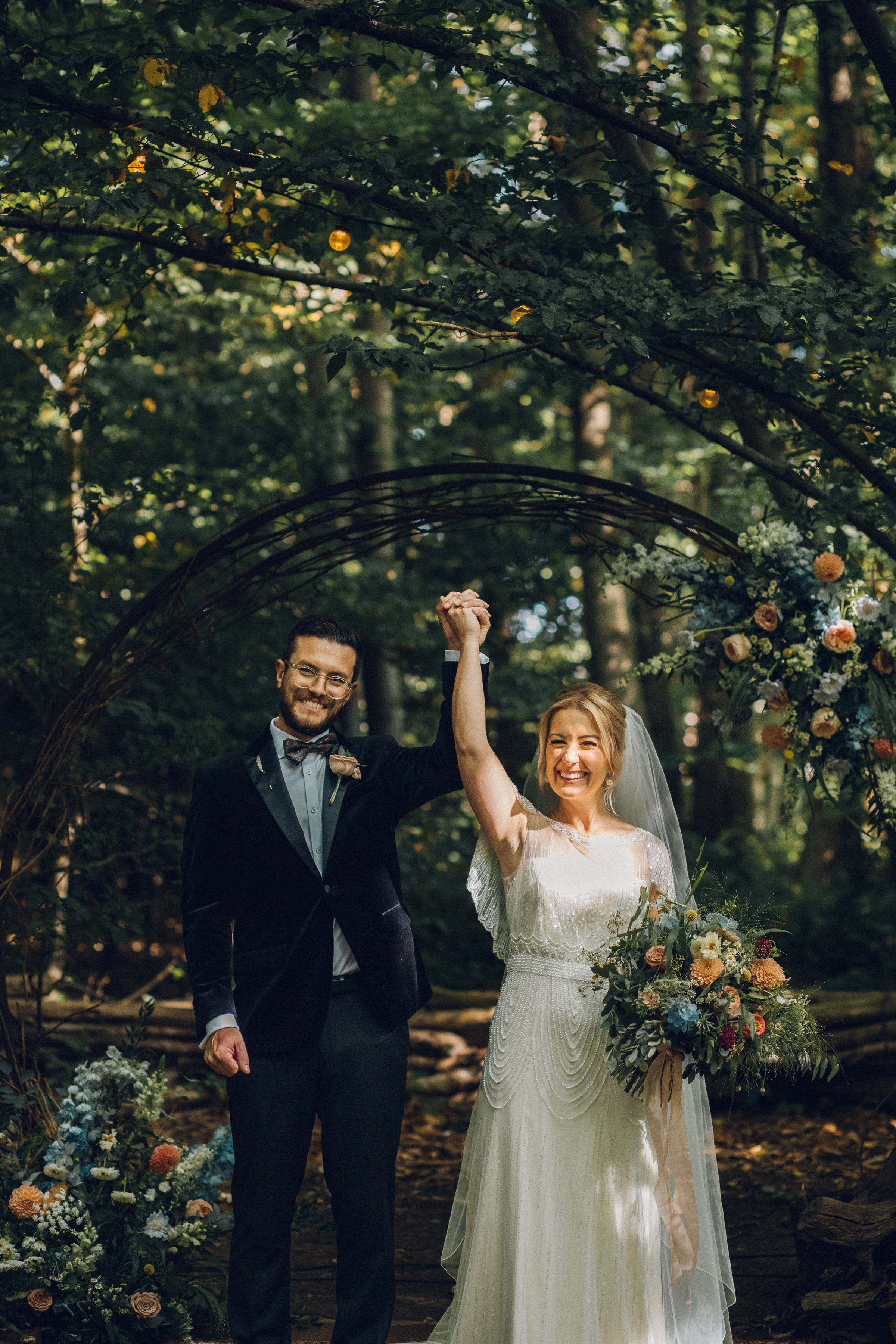 A bride and groom raise their hands together infront of a wooded area