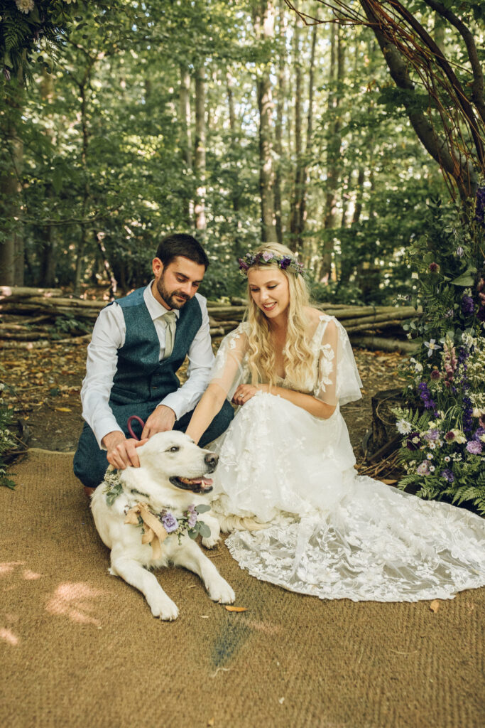 A bride and groom neal to pet their dog infront of a woodland area as they have their wedding videos filmed